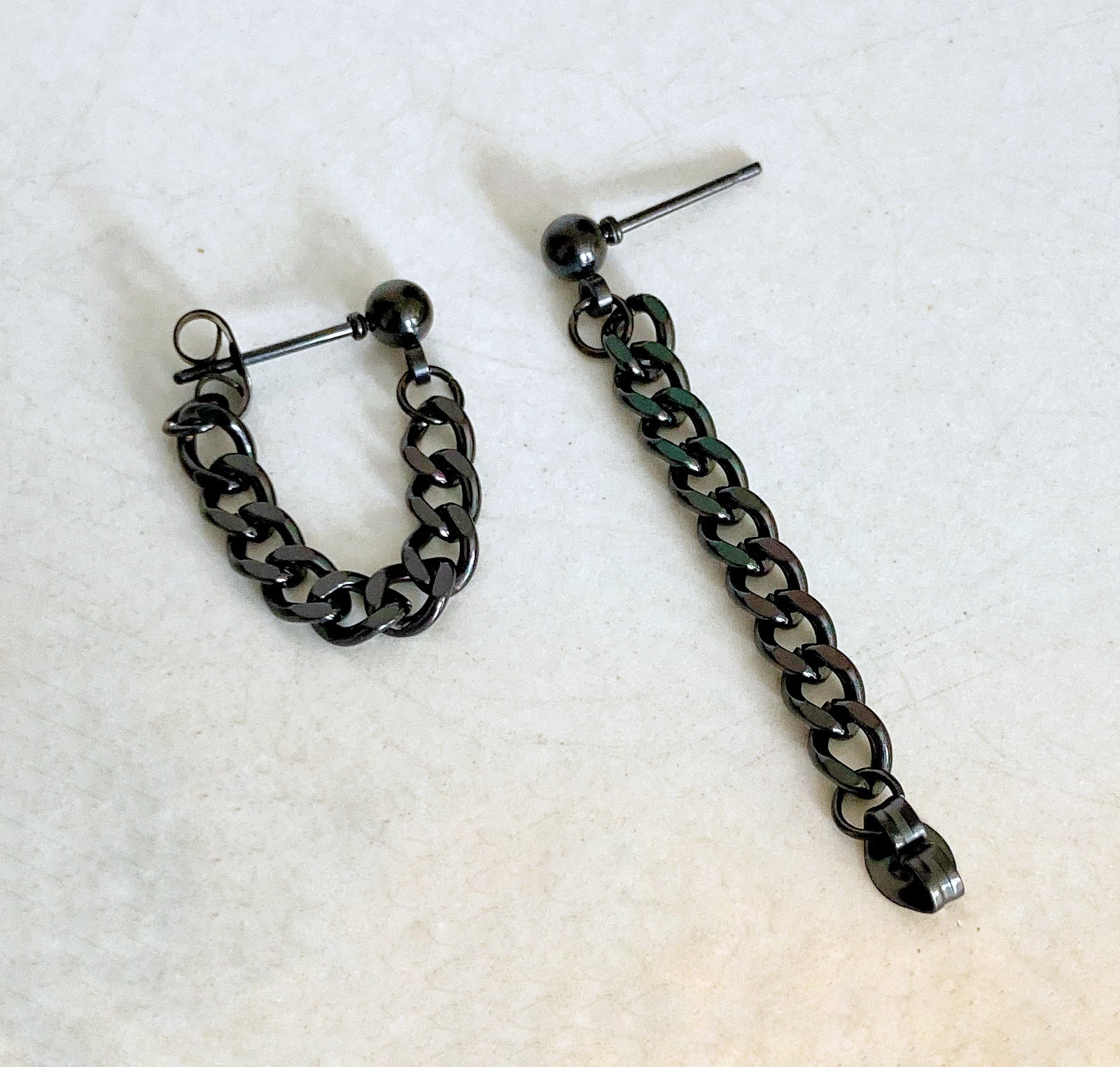 Two earrings lie next to each other. Black in colour one shows a chain lying stretched out and the other shows it fastened. The chain is attached one end to a ball shaped stud and on the other end is a butterfly backing. 