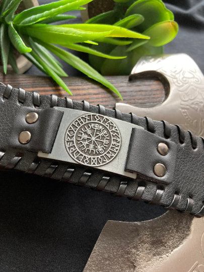 A black leather bracelet sits on the edge of a wooden axe with a steel blade and has a green plant in the background. This is a wide bracelet which has a silver coloured metal plate with a vegvisir compass engraved onto it and studs either side.