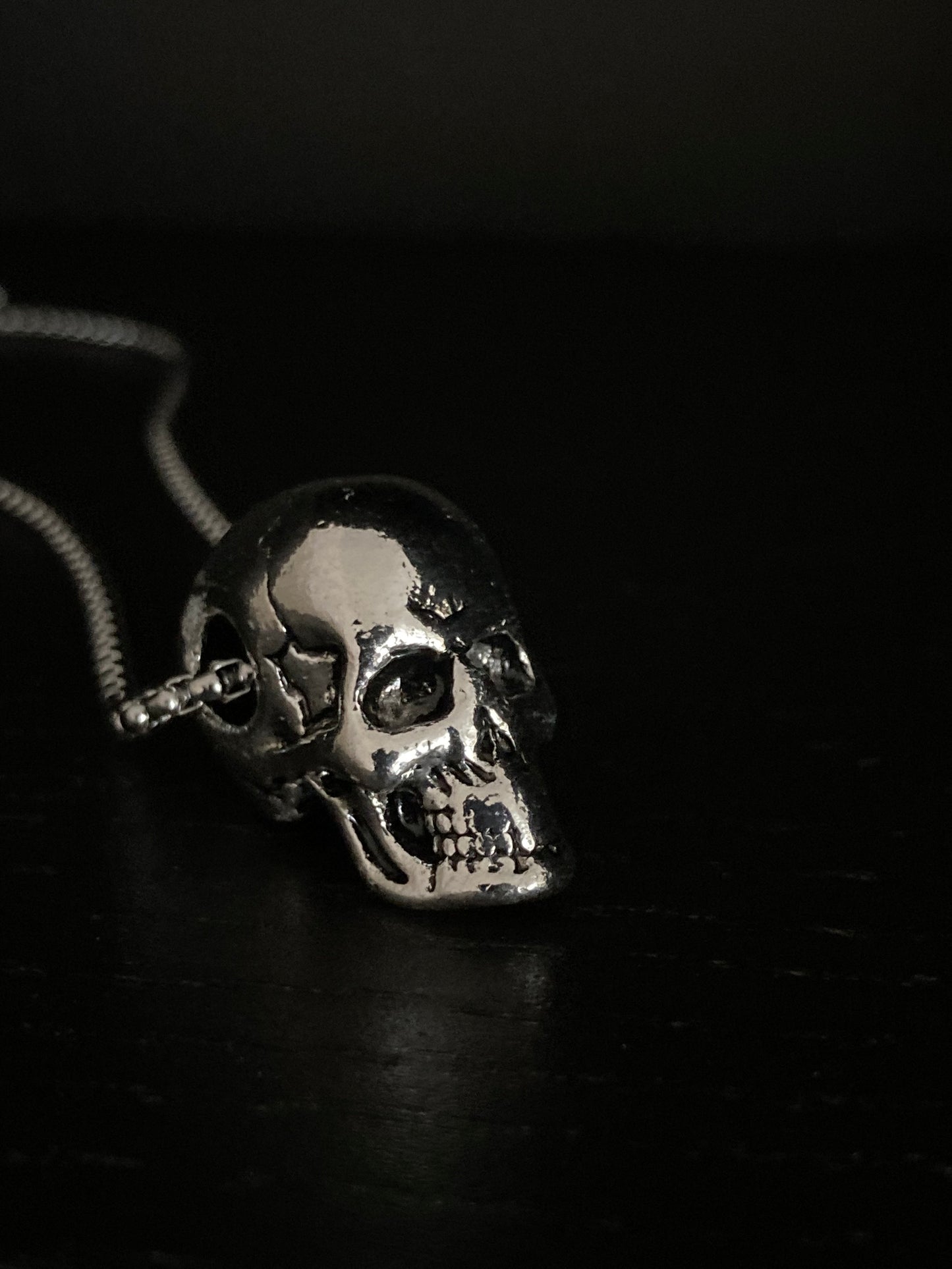 A side view of a solid skull head pendant. It is silver in  colour as is the sturdy chain and is stainless steel. The skull has detailed features including sunken eyes, a wide grimace and cracks in the skull. This is a chucky but not too weighty item