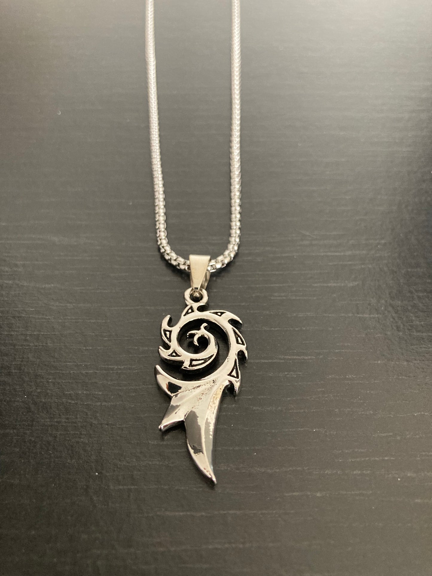 A close up view of a beautiful spiral shaped pendant which is silver in colour and made of stainless steel the same as the chain attached to it is. The bottom on the pendant is a long spike which then leadsd up to the spiral itself.