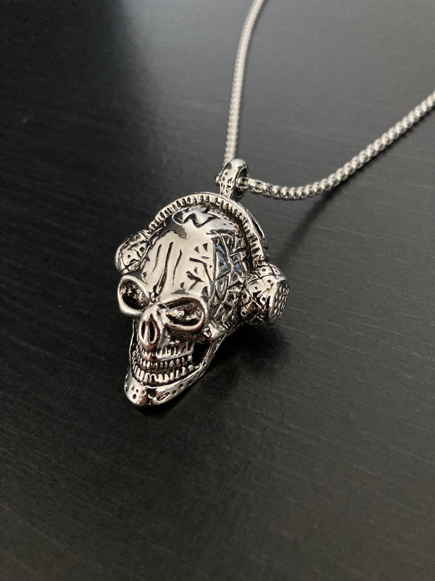 A solid looking stainless steel skull pendant sits on a black surface. It is silver in colour as is the chain attached to it. The skull is wearing a pair of headphones and has an evil grin on its face. The item is weighty to hold and very chunky.