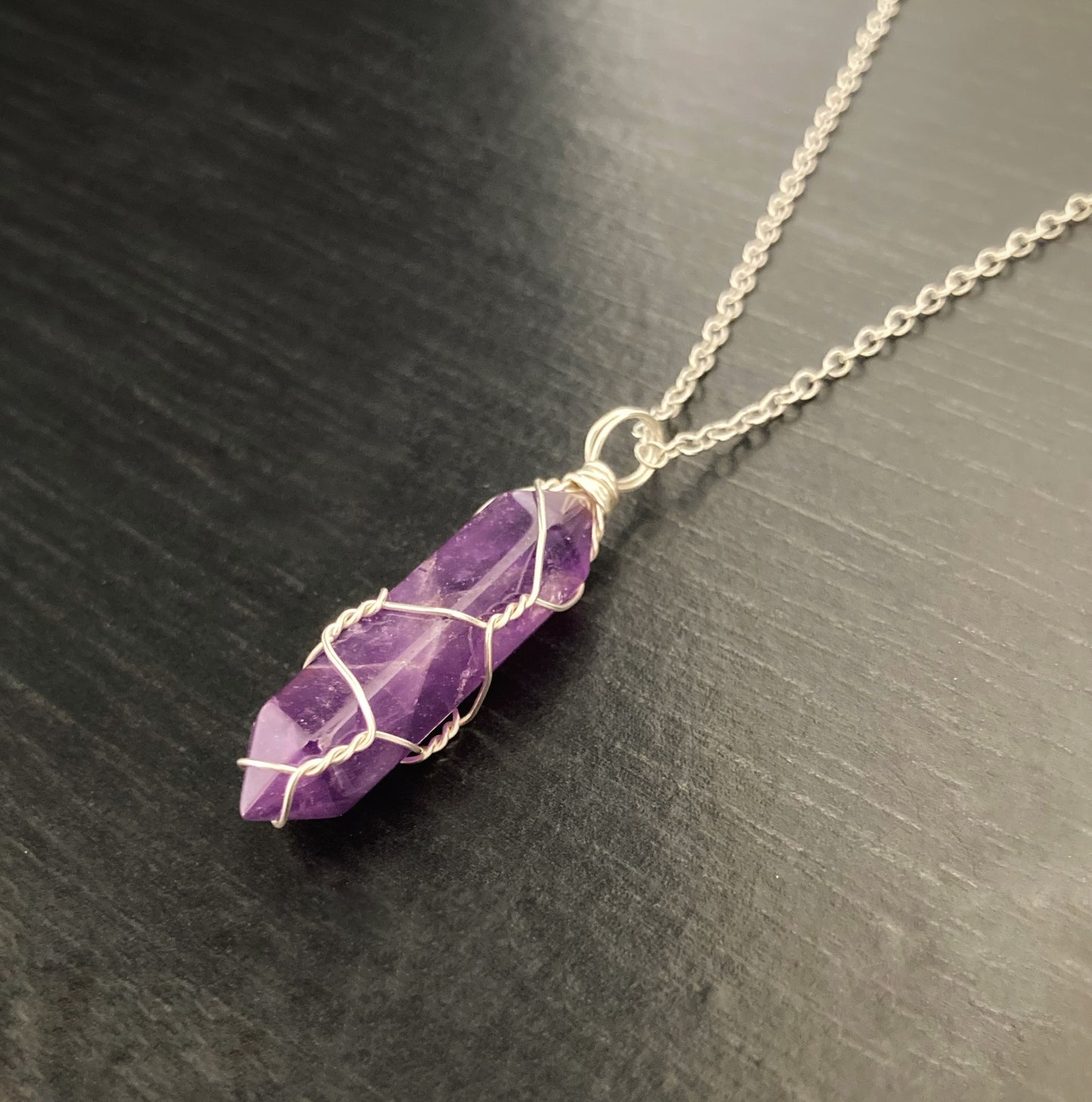 A purple coloured crystal wrapped in thin silver wire is attached to a chained necklace also silver. 