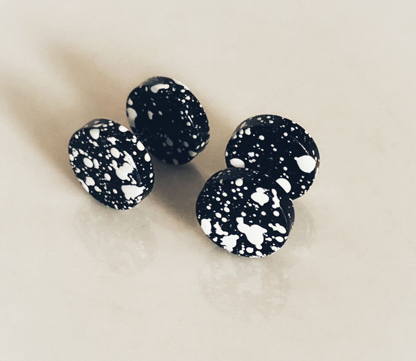 Two fake plug earrrings sit side by side on a white surface. These are black with white splodges all over the them giving the appearance of paint splatter. 