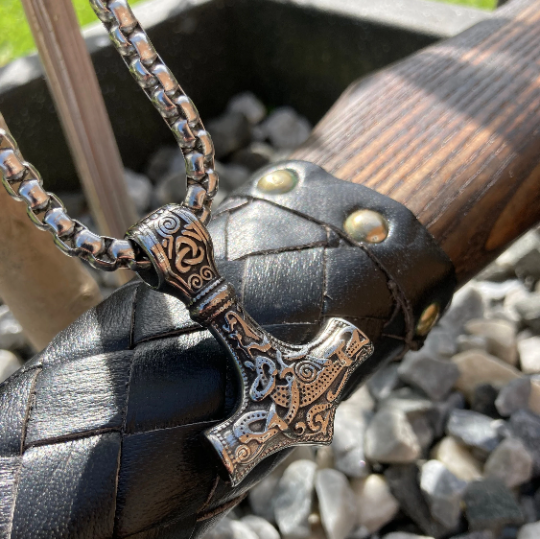 Draped across the leather part of an axe handle is a solid stainless steel silver coloured necklace in the shape of a viking hammer. Part of the chunky chain is showing and you can fully see the detail on the main body of the pendant. 