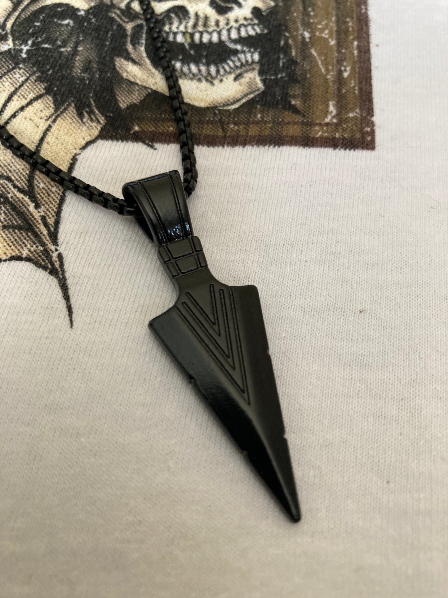 On a white background where a skull drawing can be seen is a close up view of an arrow head necklace. The item and the chain are both black and it is lightweight to hold. There are v shaped lines engraved onto the arrow itself giving it a cool look.