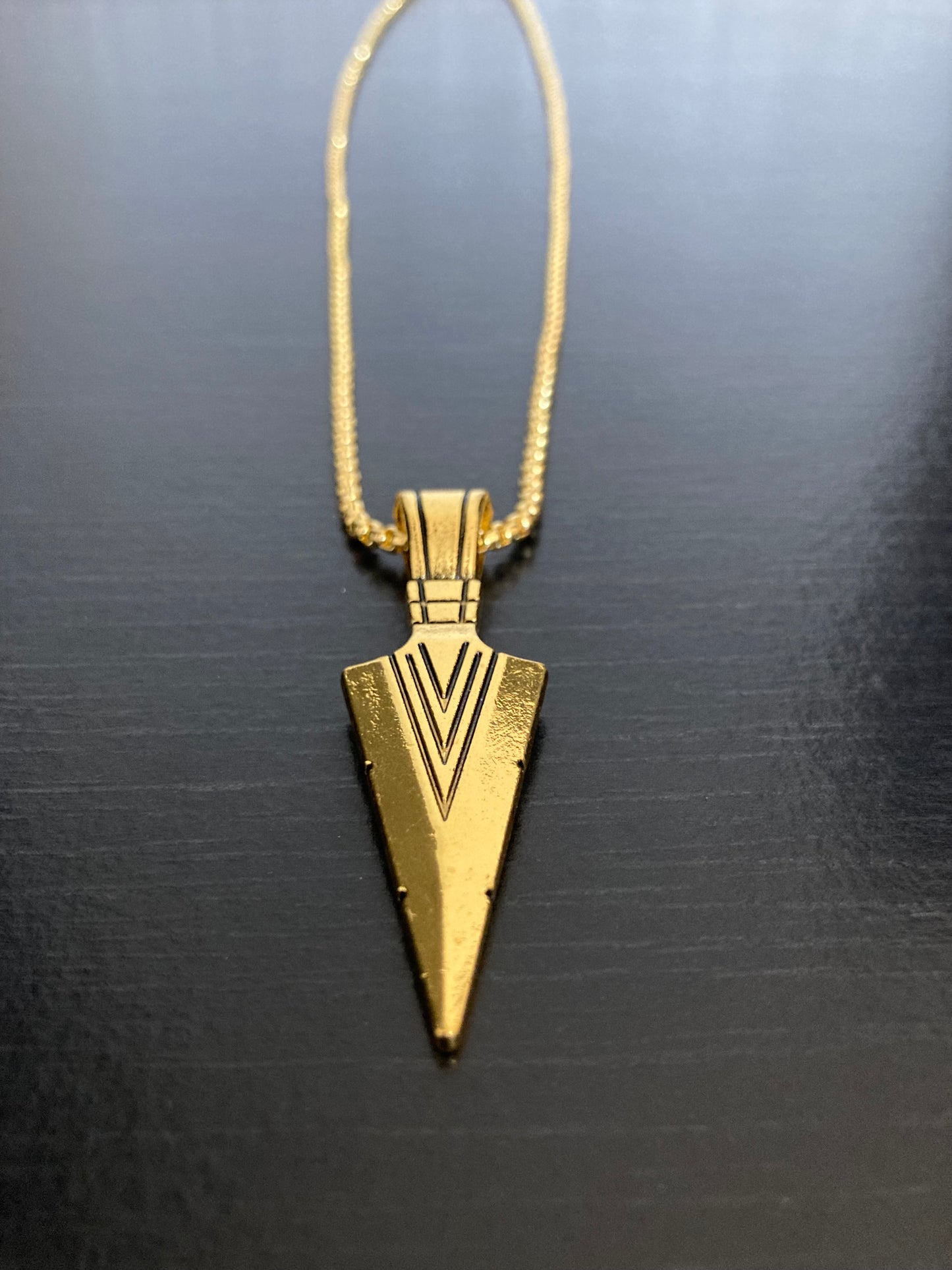 A grainy black surface holds a arrow head shaped necklace. The pendant and chain are both gold in colour and made from quality stainless steel. There are markings on the arrow which you can feel when you touch it.