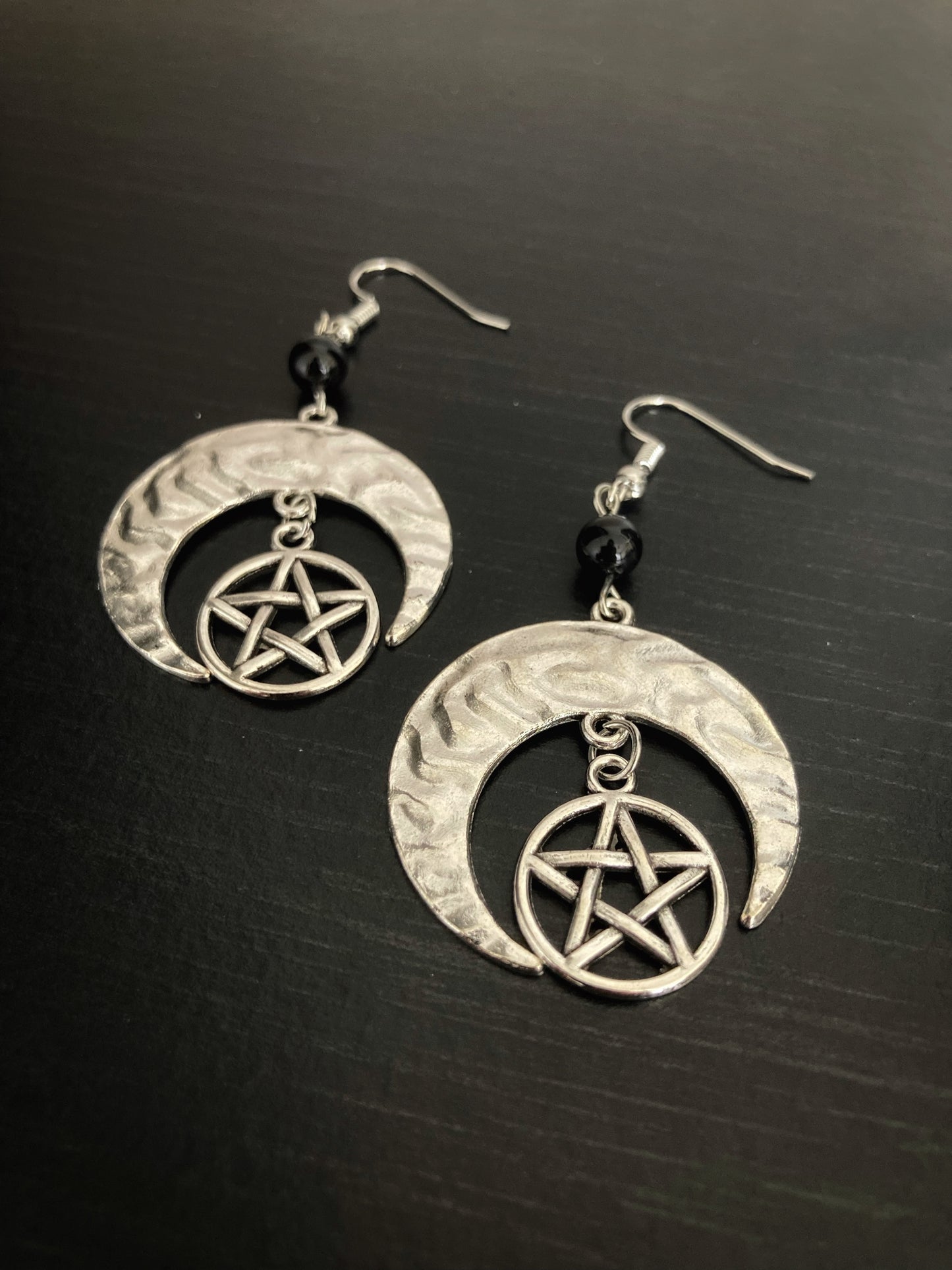 Earrings lie on a black background. There is a pentagram on each one that hangs from the inner part of a crescent moon shape. They are a shiny silver colour and both feature a black round bead above the moon. The surface of the crescents are slightly rippling in texture.