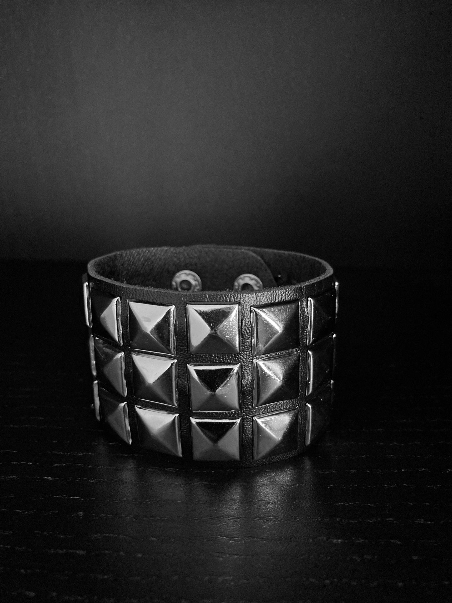 A wide cuff bracelet is on view. It is black and leather and on the outside are three dimensional pyramid shapes within small square pieces. The square bits arer three across the width of the bracelet and are shiny in appearance.