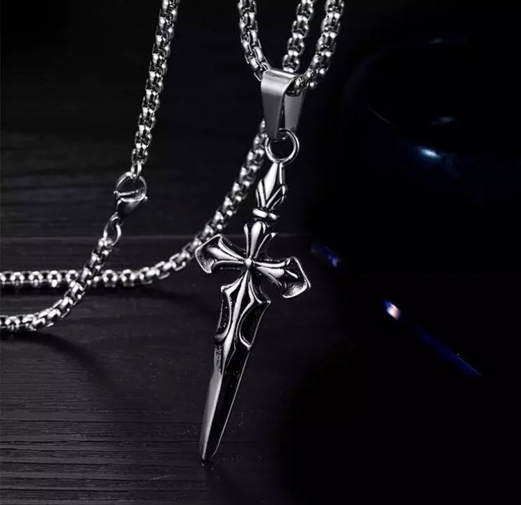 The point of a sword pendant rests on a black surface. You can clearly see the chain its hangs from and the clasp that fastens the necklace. There is a shine to the whole thing and is silver in colour.