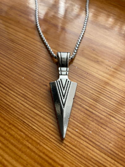 Upon a varnished wooden surface sits a small arrow head necklace. There is a chain attached to it which is of good quality and is sturdy. The whole thing is silver in colour with a shiny appearance and textured lines engraved onto the arrow.