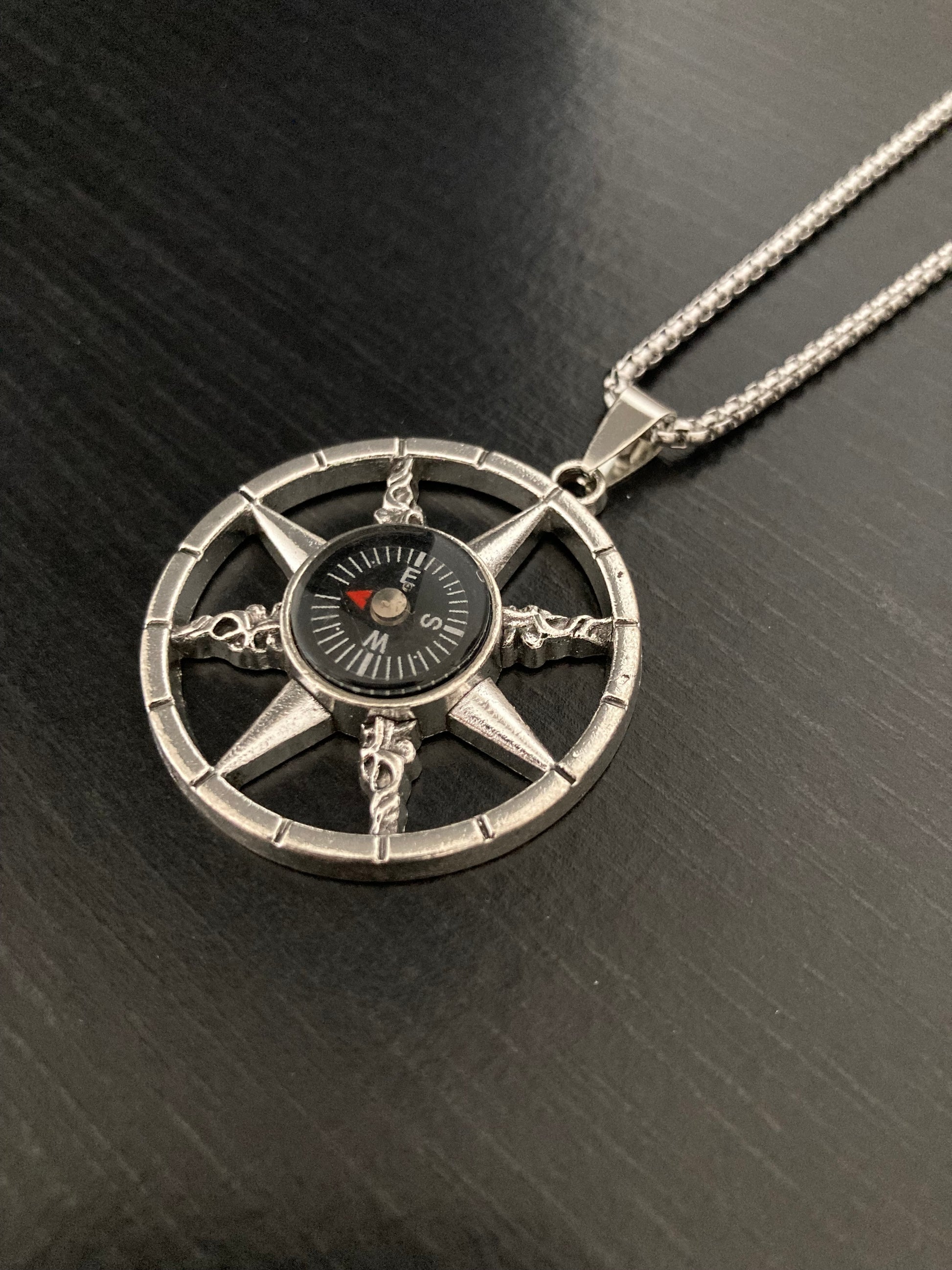 Lying angled on a black surface is a fully working compass necklace. Attached to a stainless steel chain the pendant is a circle shape with the compass in the middle of it. Around the centre are cut out points which are against the outer ring.