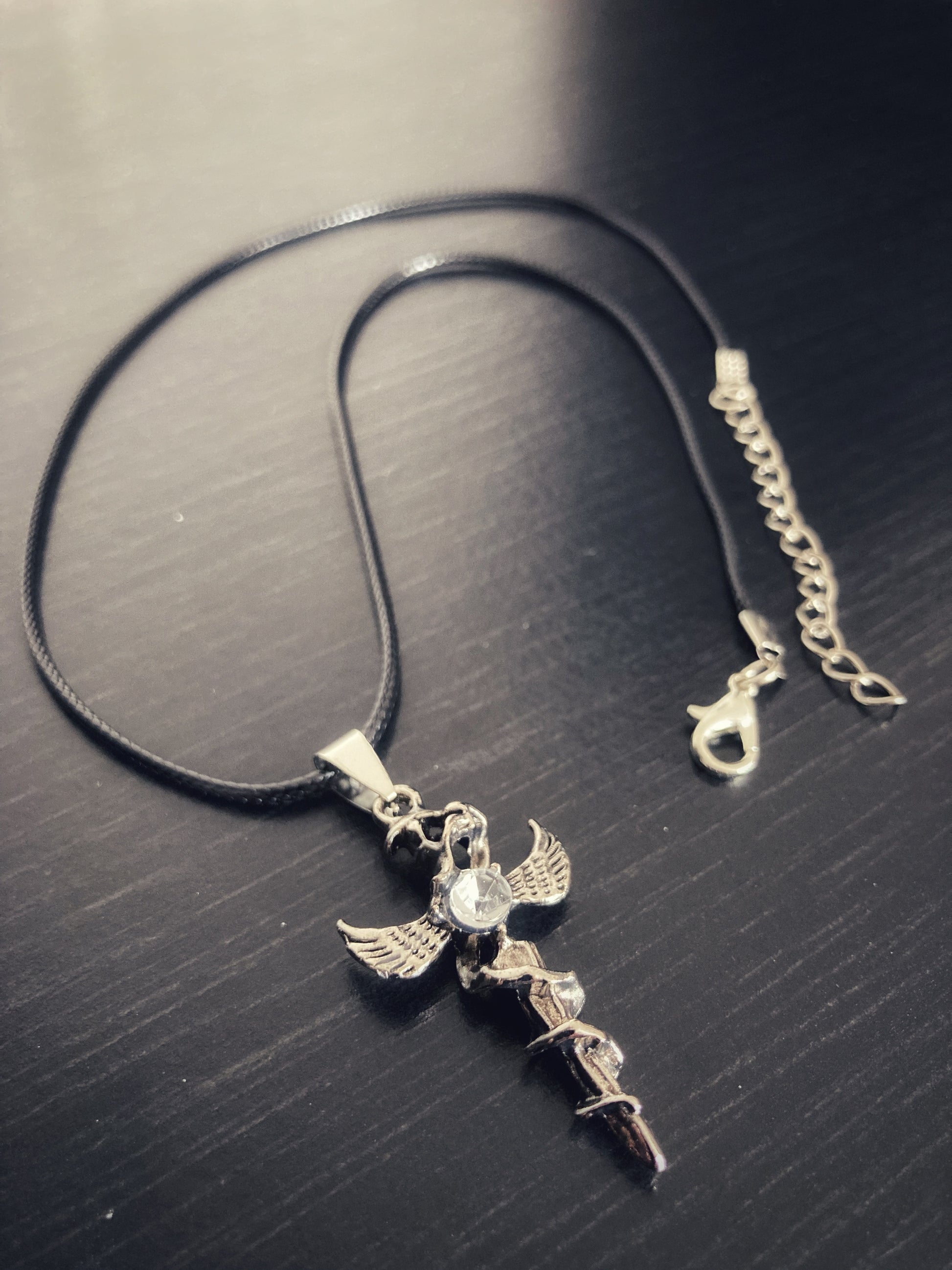 A full view of a black corded necklace with metal clasp attached to a dagger shaped pendant. There is a winged serpant which is wrapped around the dagger and a clear glass stone is attached to this. It is light in weight and slightly chunky. 