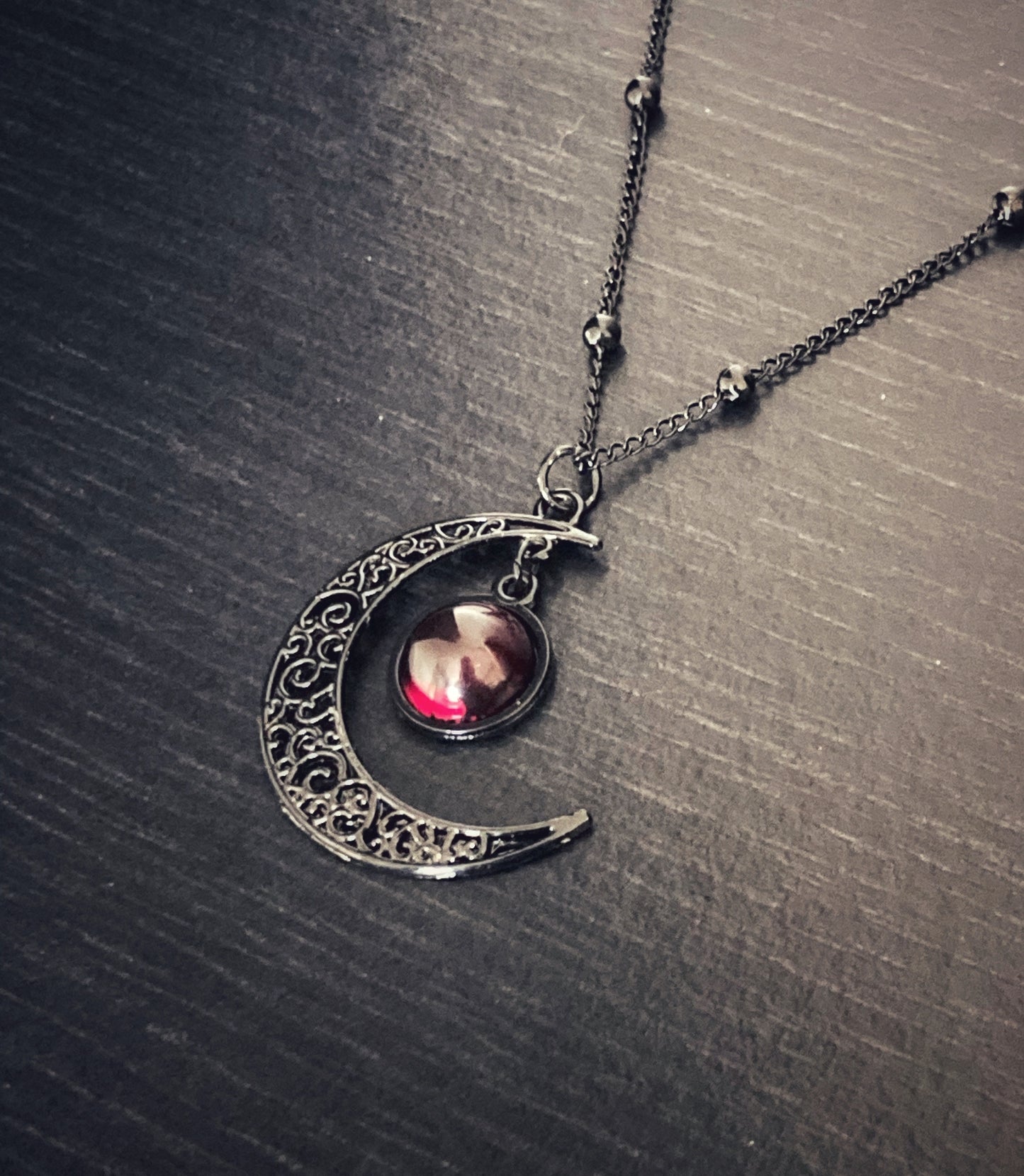 A gothic looking crescent moon pendant is sitting at an angle. You can see the narrow link chain attached to it and both are black. There is a dome shaped red glass looking stone hanging from the moon into the curve that glistens in the light.