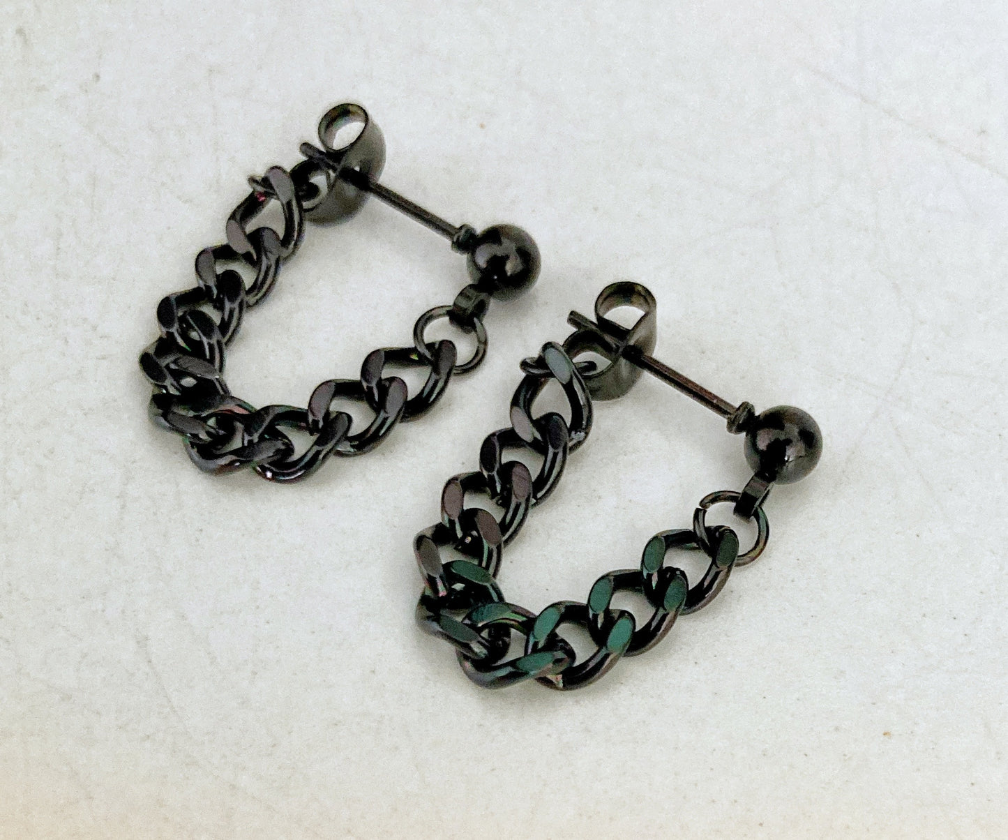 A pair of black stud earrings sit side by side. There is a link chain that is attached to the stud with the other end being a butterfly back, creating a loooped chain. Shiny in appearance and lightweight to hold.