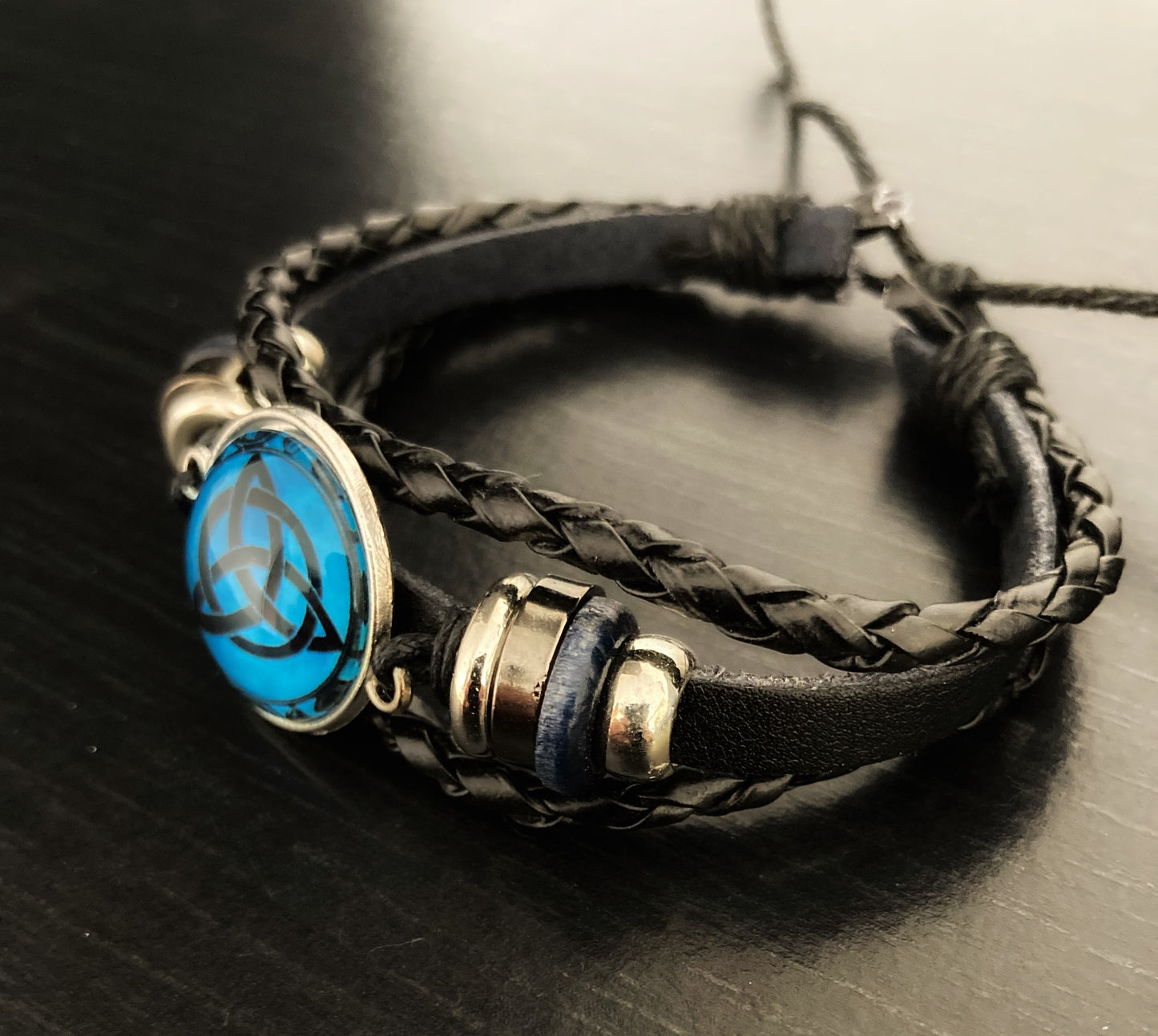 A black leather bracelet sits on a dark surface, this is made up of 3 bands that are all tied with a knot to keep them together. This fastening is adustable to fit many sizes of wrist.