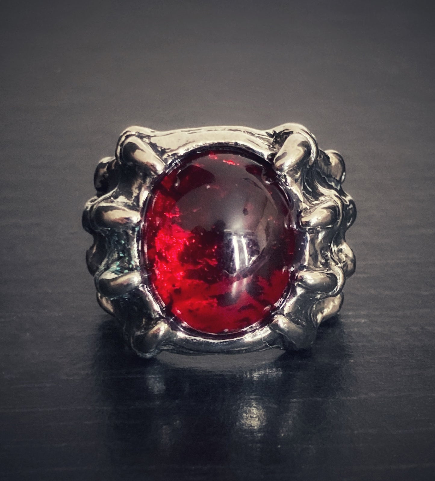 A silver coloured ring sits on a grainy black surface. This is a front view of an item which is made to look like a dragon claw that is holding onto a red stone which is made of glass. When you hold the ring you can feel the detailed markings on it.