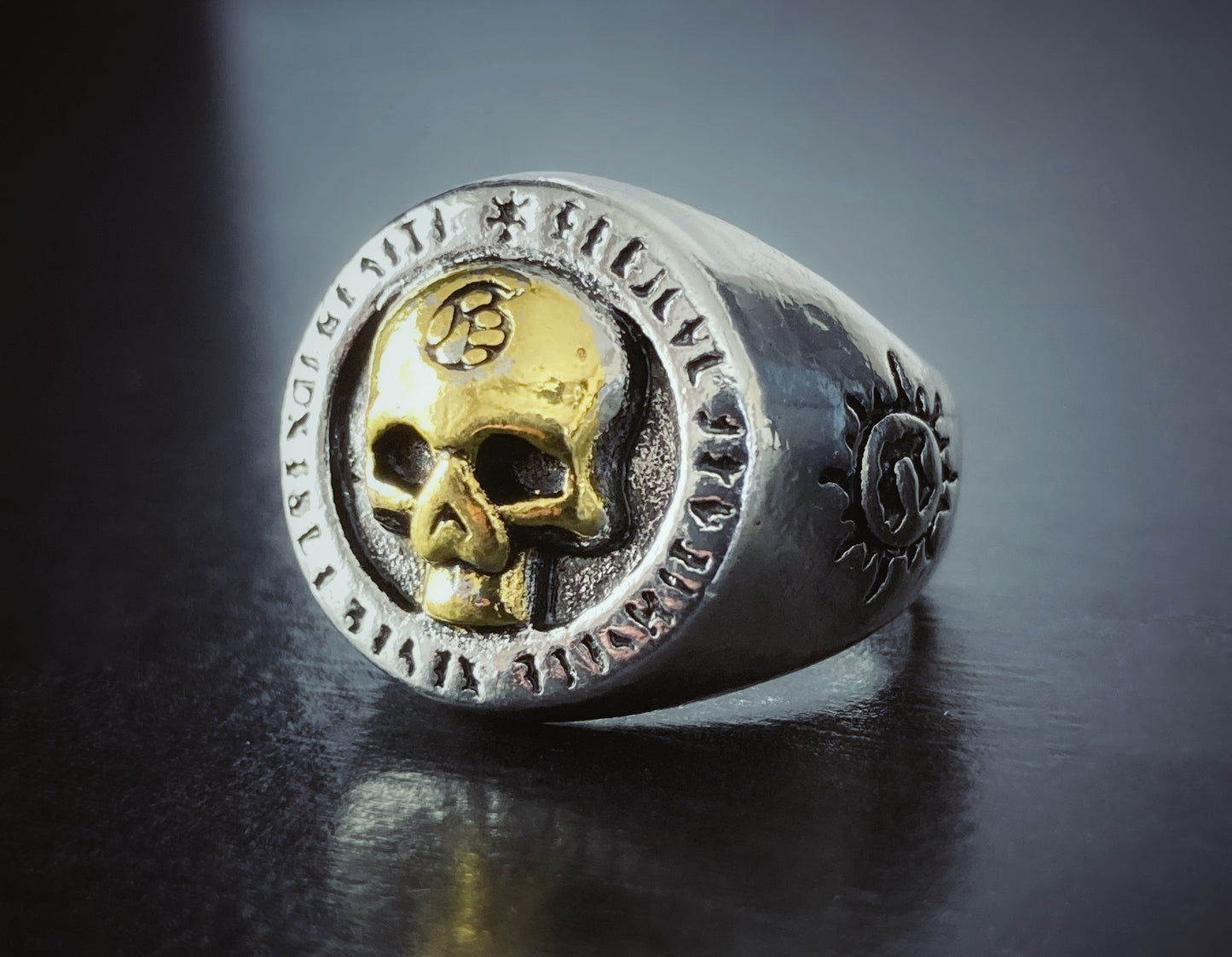 A biker style stainless steel ring sits on a black surface. It is fairly shiny and has a skull engraved onto the top part of it. 