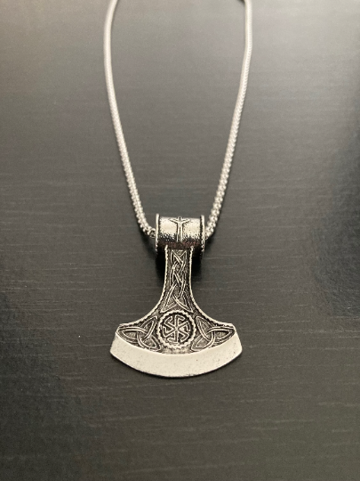 The head of an axe makes up the pendant of this item which is sitting on a plain black background with a chain attached to it. They are both silver in colour and made of solid stainless steel. 