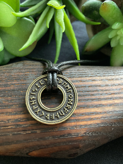 Lying draped over a wooded surface is a circular pendant with the middle cut out. It is a gold colour with runic letters that go around it. It is very light to hold and a corded necklace is tied to it.