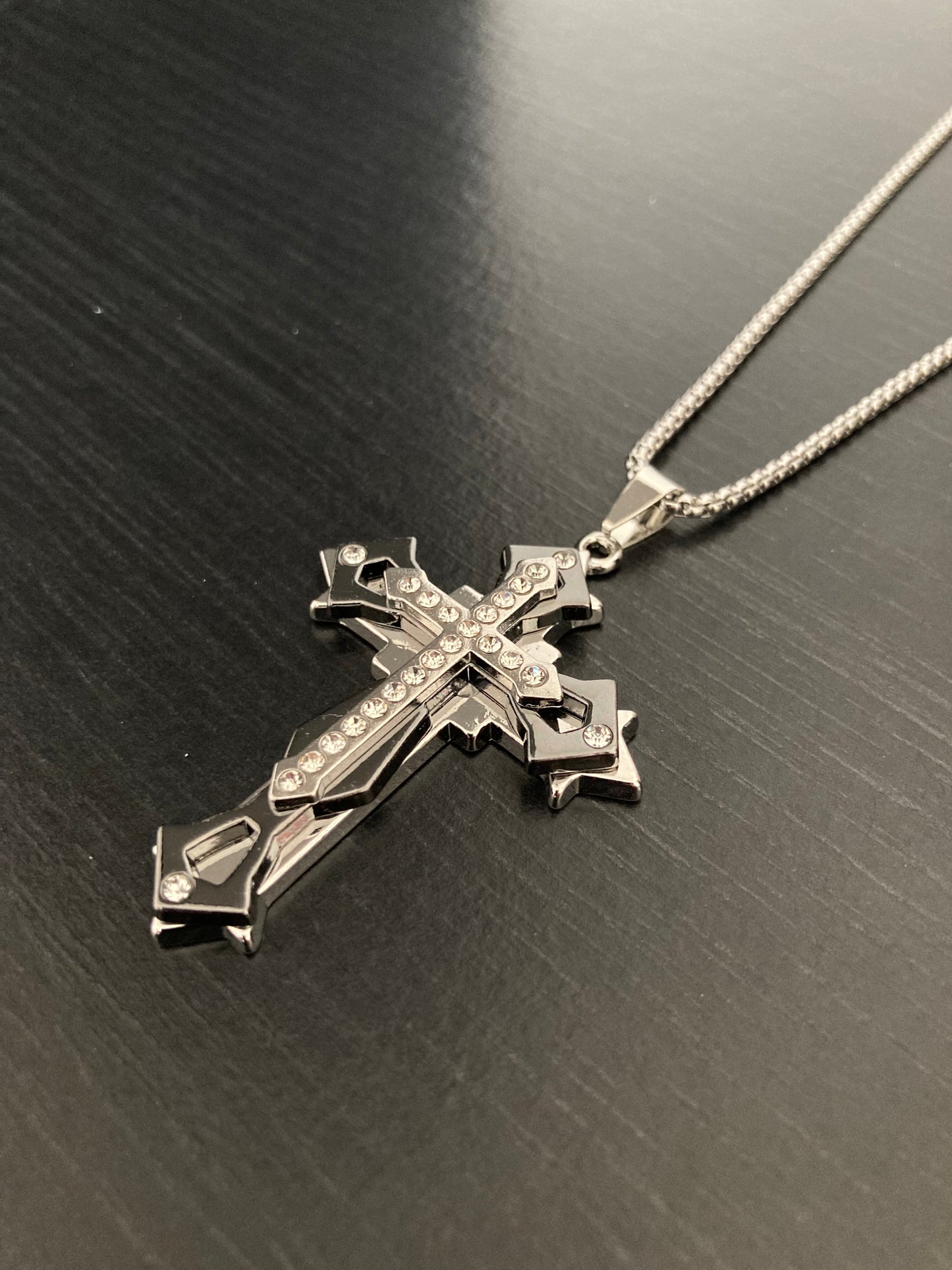 An angled view of a solid looking cross pendant on a chain. Silver in colour there are 3 crosses of varying sizes atop each other. The top cross has a bejewelled design that runs down the length of it.