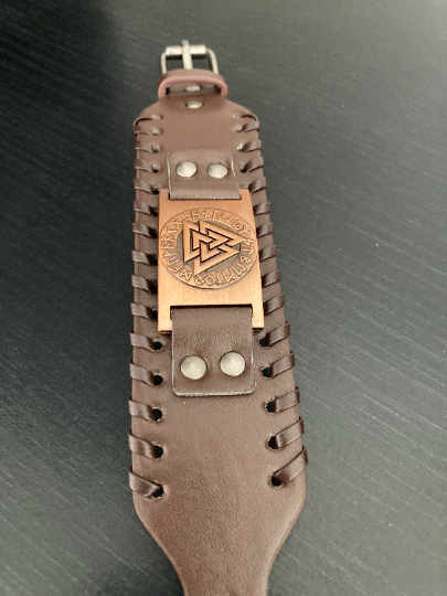 A wide leather bracelet sits stretched out on a plain black background. The item is brown in colour and features a bronze colour metal plate as the main design which has the viking valknut surrounded by runes engraved onto it. 