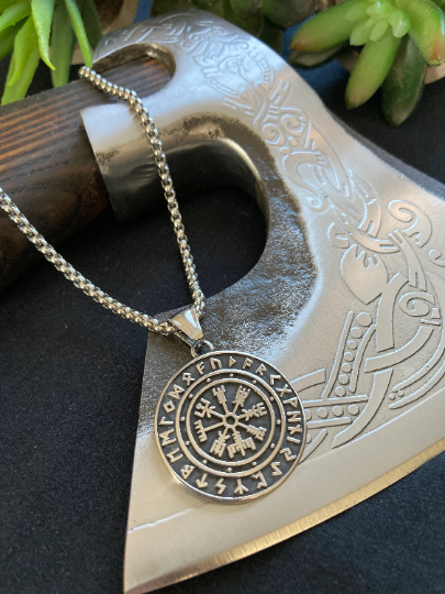 A wooden axe handle is featured with the necklace lying over it on a black background. The main focus is on the compass design which is a raised design which you can feel when touching it and this stands out on the background of the pendant.