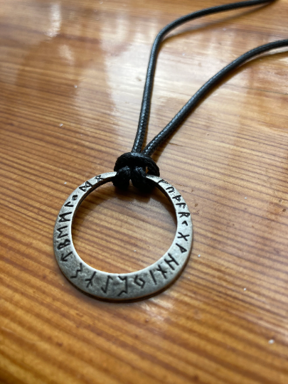 A shiny brown wooden table holds a silver coloured circular ring pendant which is hollow in the centre and has rune letters running around the edge of it. Draped behind it is the black cord that is the necklace and this is tied to the ring.