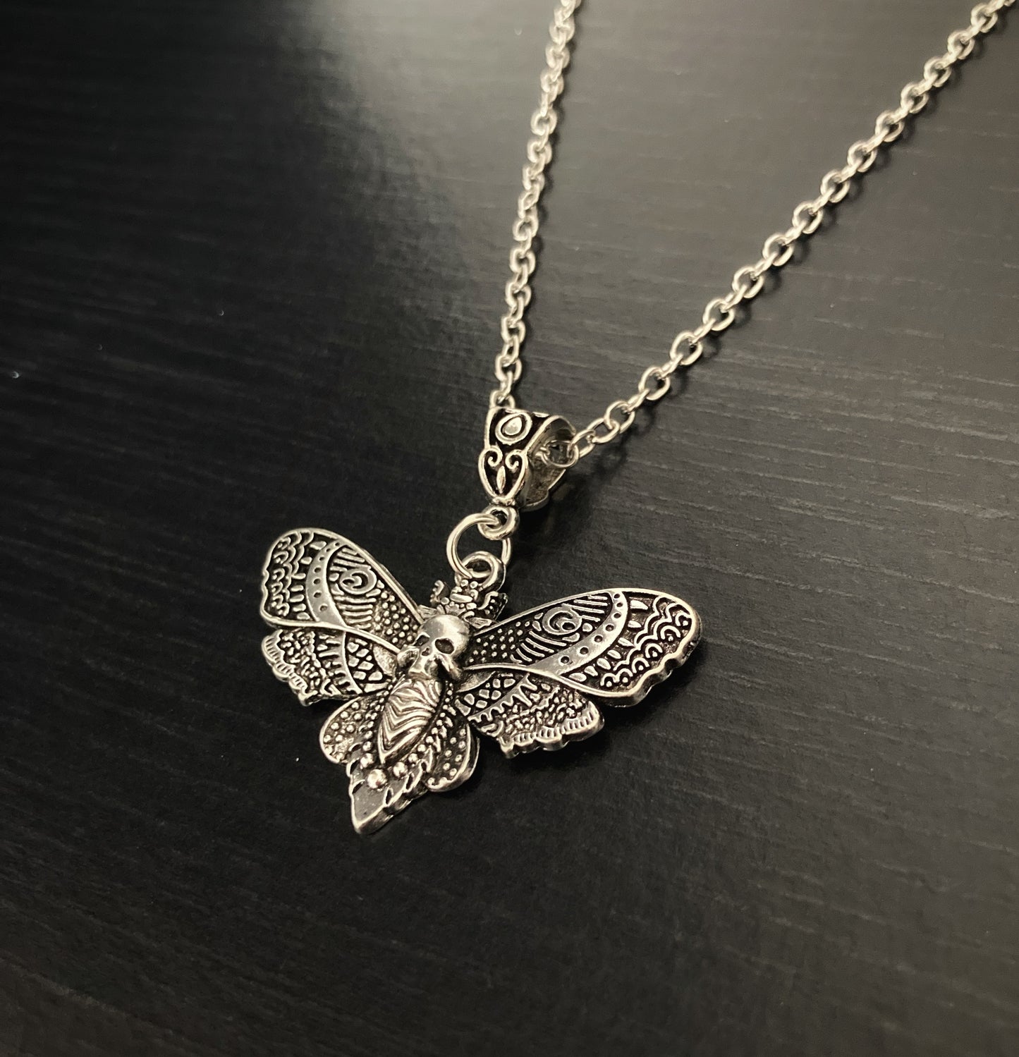 An angled view of a chain and pendant in the shape of a death moth. Made from stainless steel and silver in colour this sits on a black background and you can really see the detailing on the wings which are stunning to look at. 