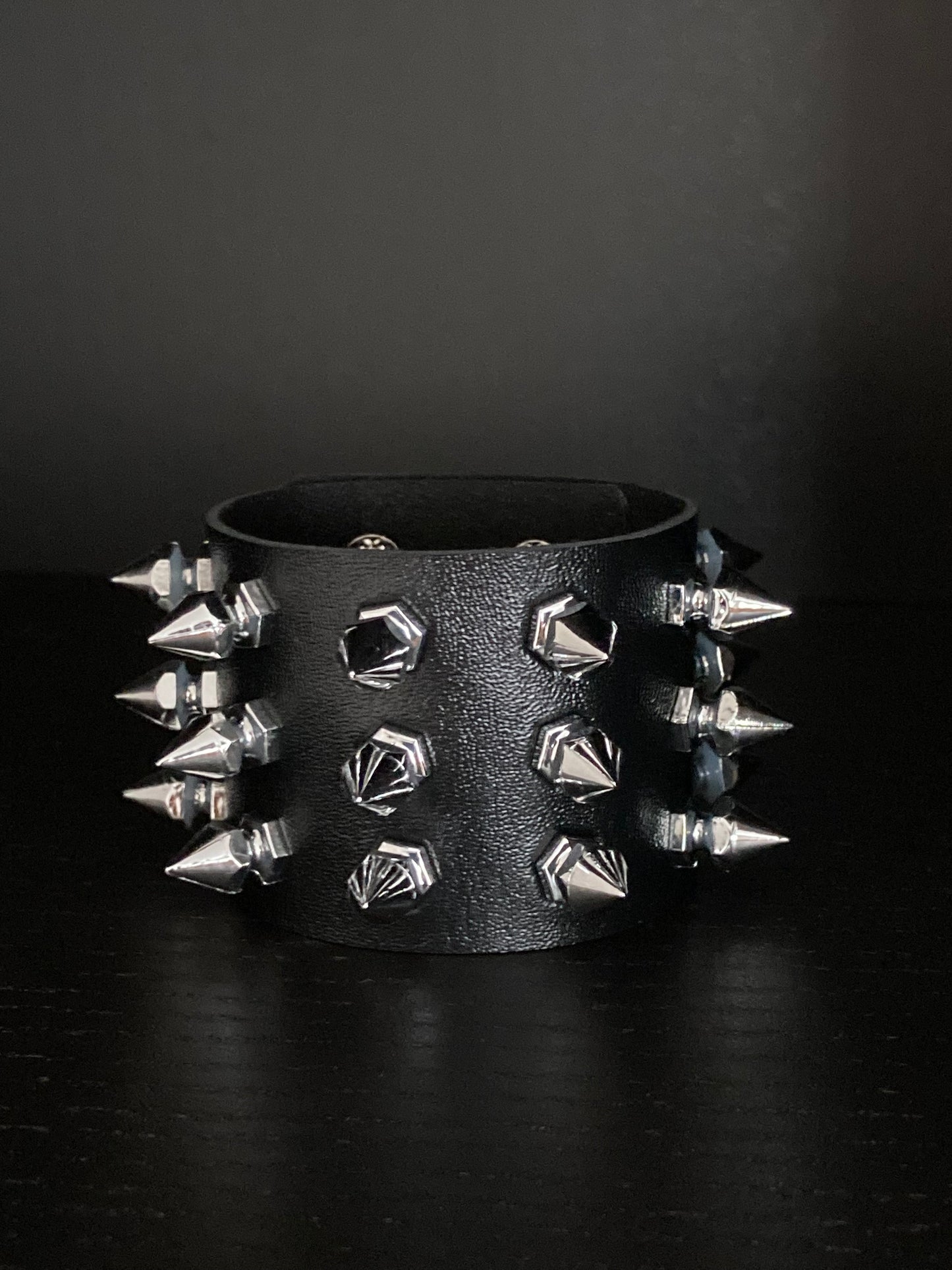 A shiny loooking black wide cuff bracelet is in view. Leather in material with silver coloured spikes sticking out of it. 