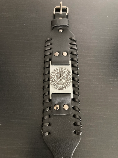 Lying on a grainy wood black background is a leather bracelet that is black and is wide. The item is laying at full stretch so you can see the length of it and the full detail including the silver colour metal buckle and the viking compass design.