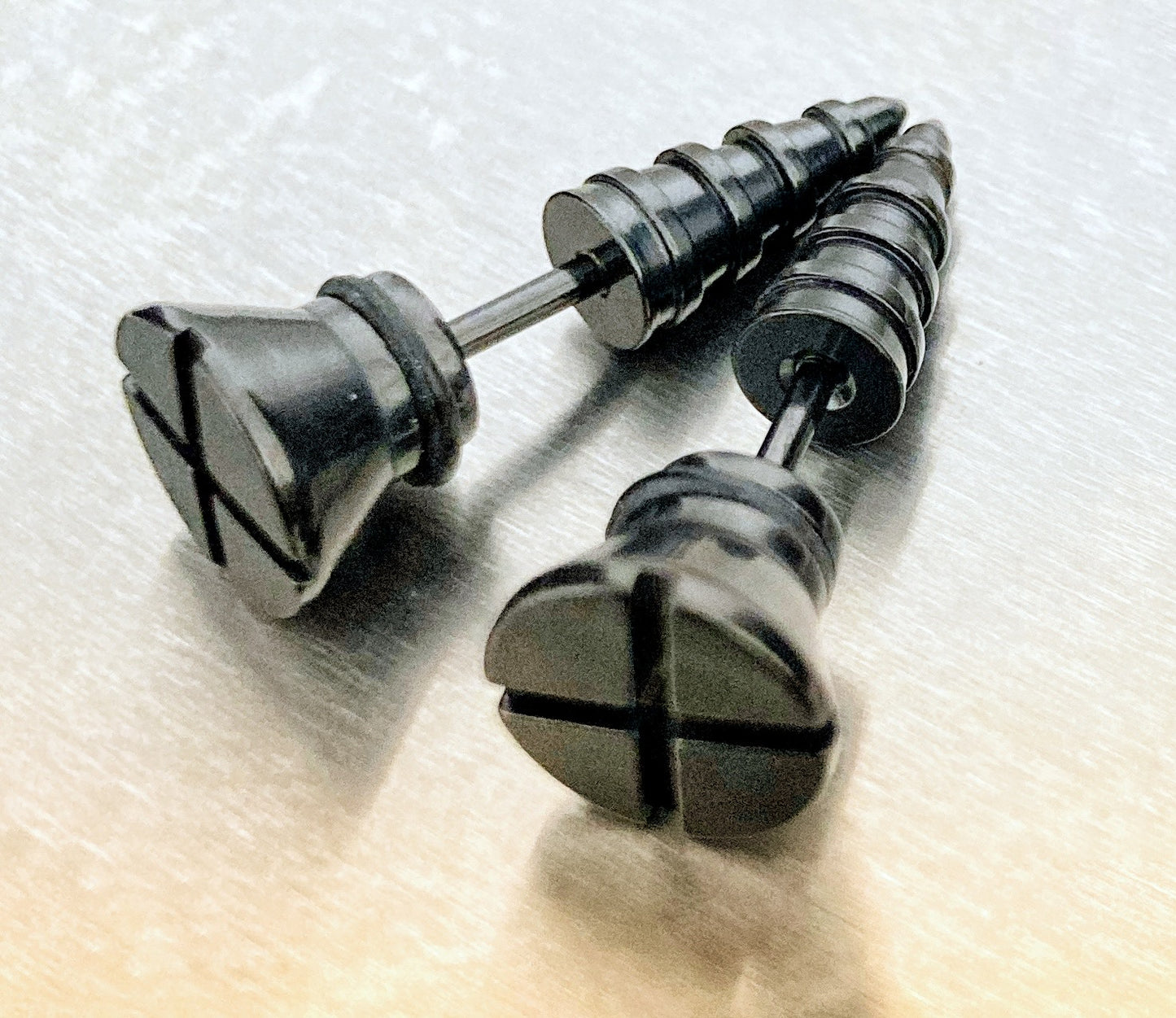 A grainy background holds a black pair of earrings in the shape of screws. The bottom halfs go down into a point and the top has a crosshead look to it just like a real screw.