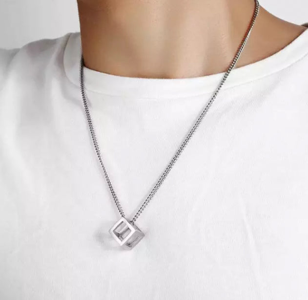 Around someones neck sits a cube necklace. This is on a chain and both are silver coloured and shiny. The cube itself is hollow not solid and it is light to wear.