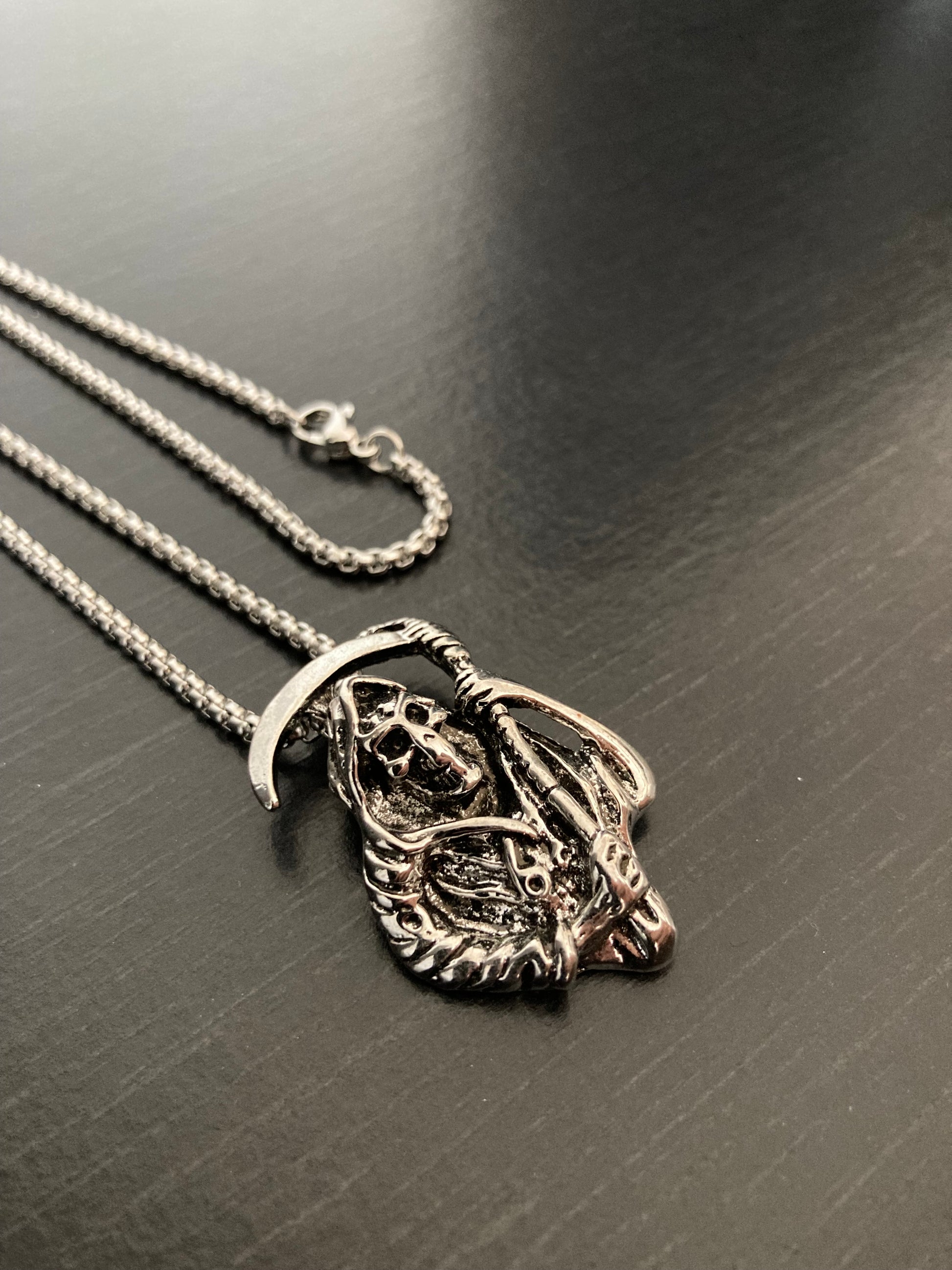 An angled view of a grim reaper pendant where you can see the quality chain and metal clasp as well as the chunky pendant. Detailed and unique looking there are lots of details to this piece including scythe, sunken eyes and a cloak. 