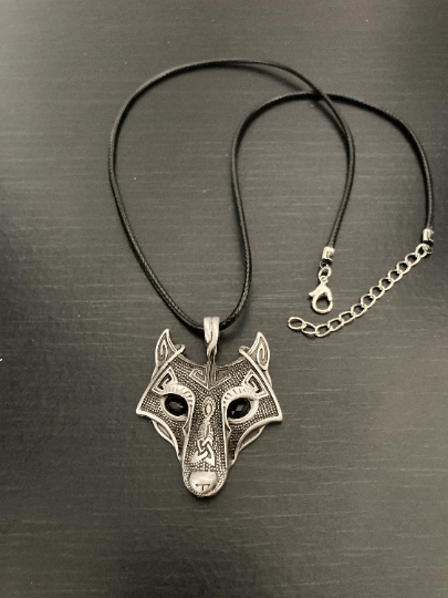 A full view of a necklace with a wolf pendant  attached to a black cord with a metal clasp. On a black background this silver coloured item is both shiny and very detailed with black glass eyes.  