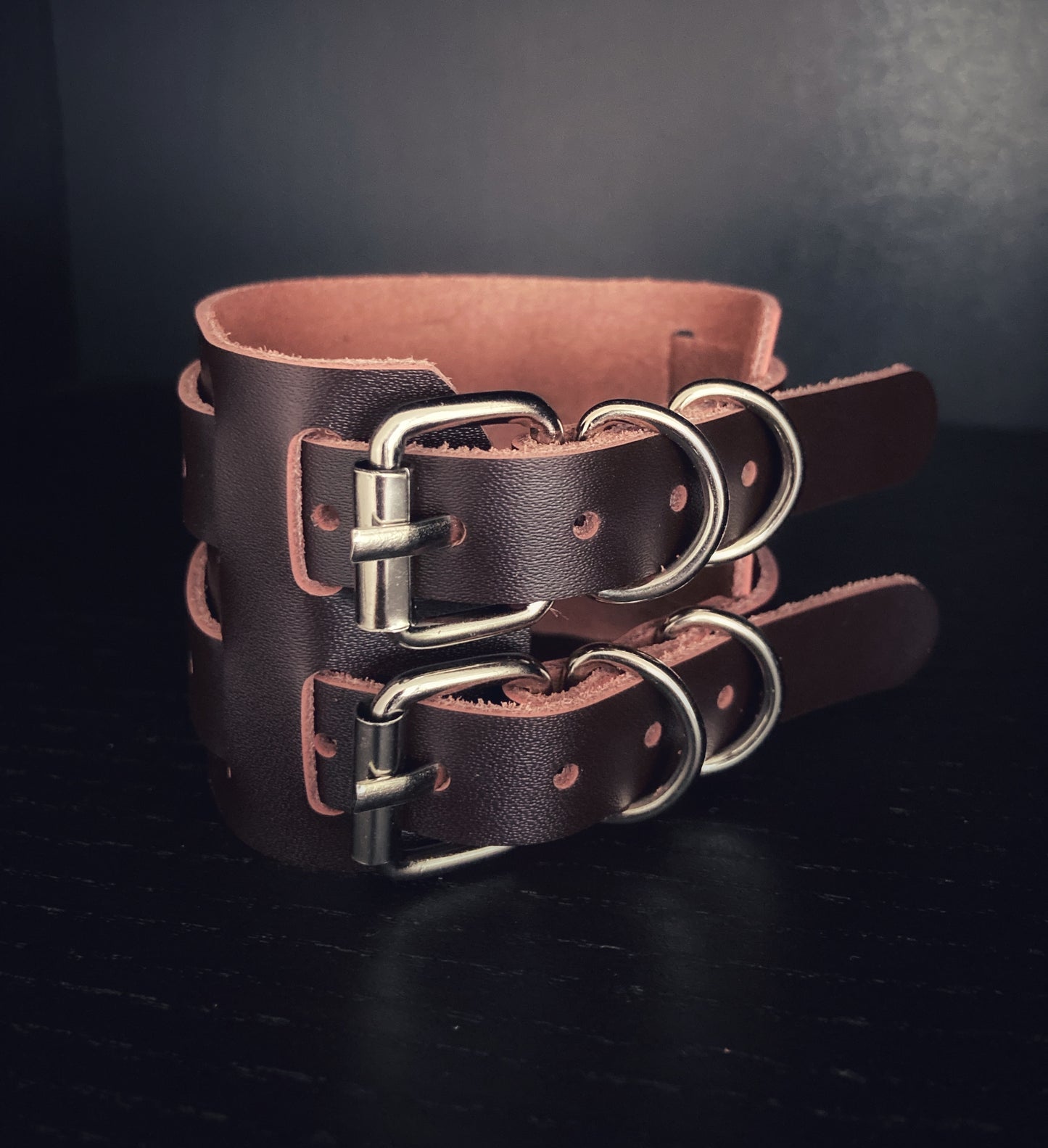 A view of two metal buckles attached to leather narrow straps. These straps are then attached to a wide cuff style brown leather bracelet.