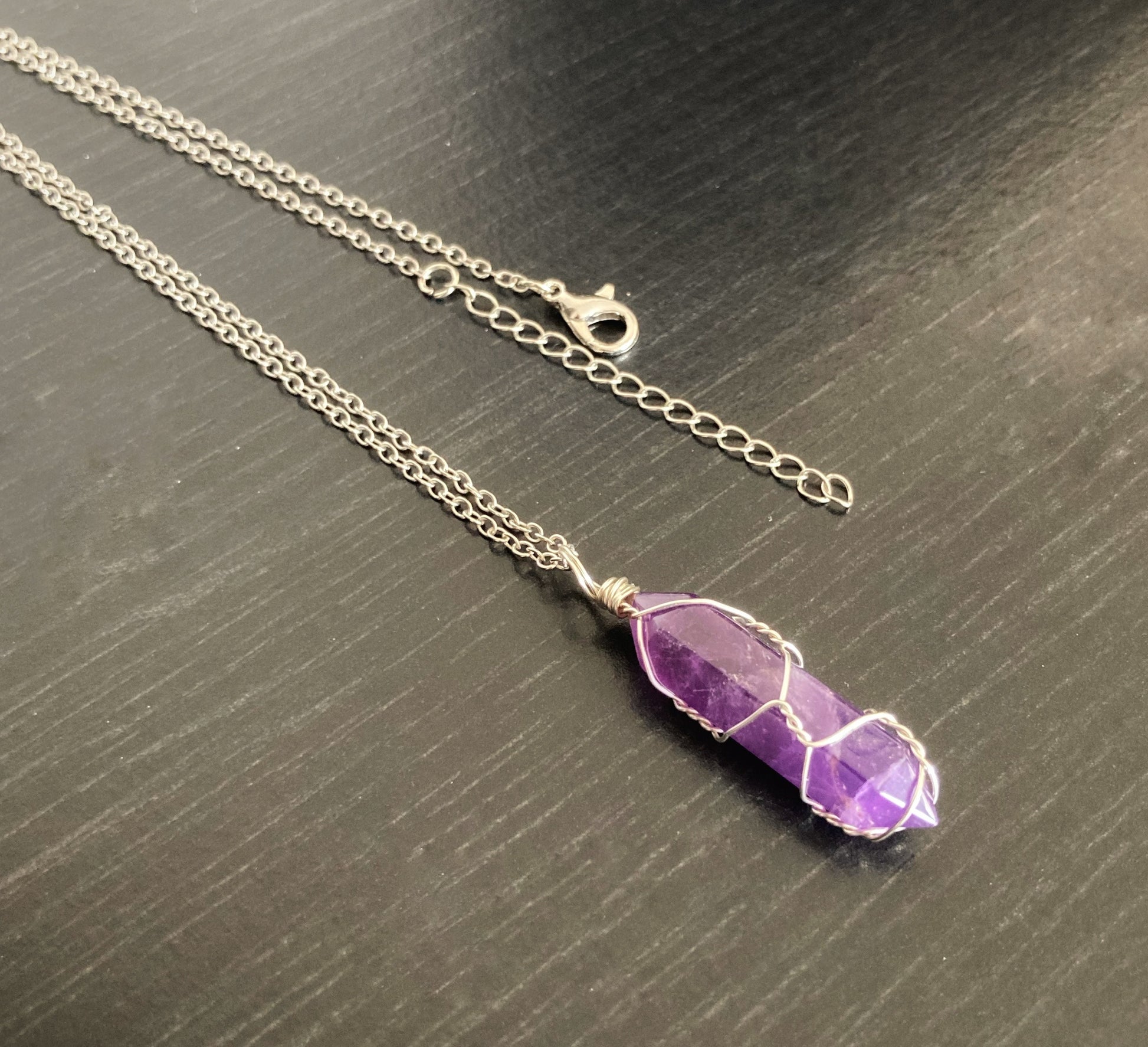 A silver coloured chain with a metal fastening clasp is on show. Lying on a black surface there is a pendant attached to it which is a purple crystal.