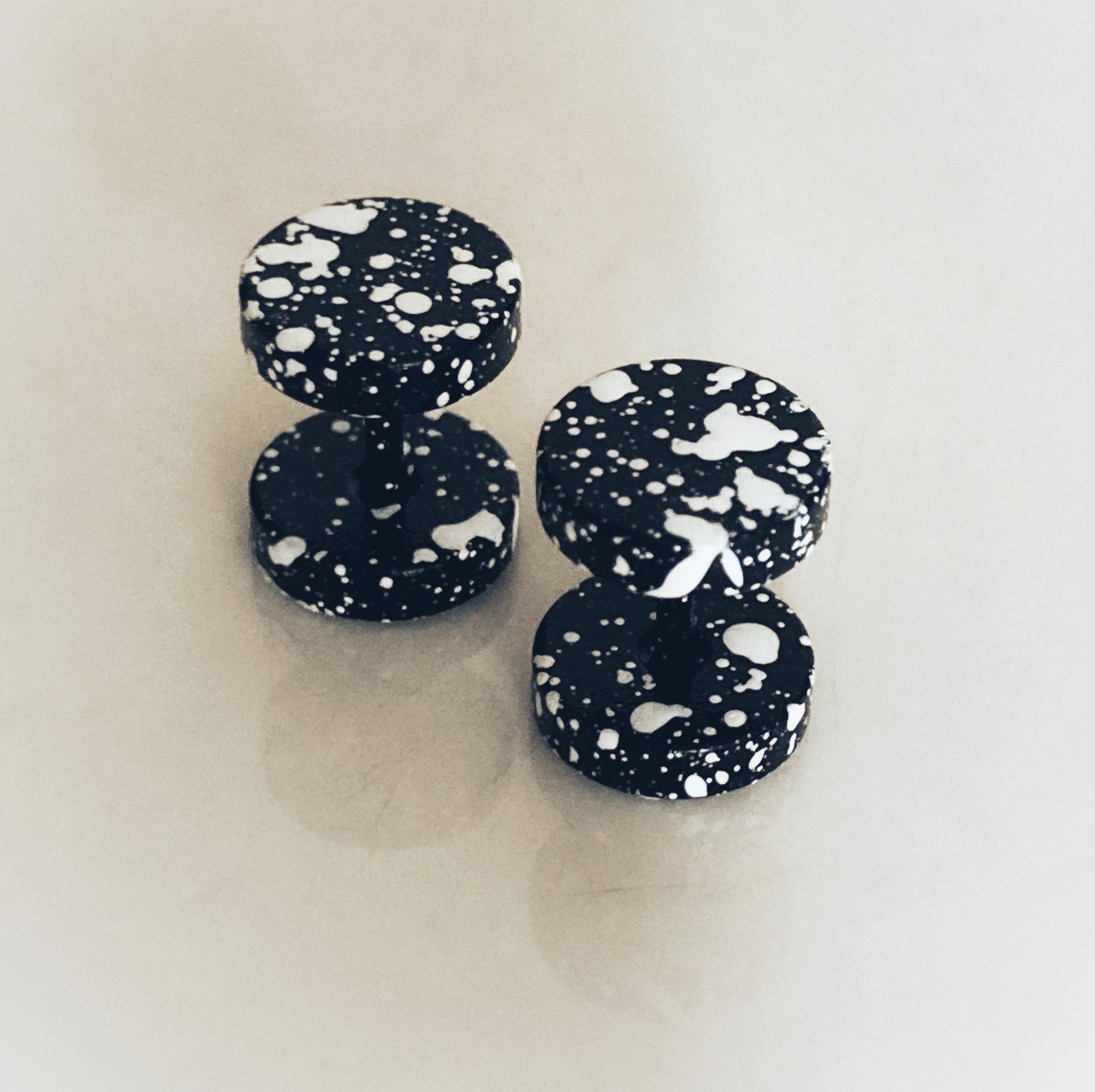 A pair of fake plug earrings sit upright next to each other. They are black with white colour paint style splatter all over them. 