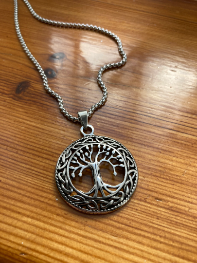 A stainless steel necklace sits on a wooden table, the main feature of this is the tree of life pendant that is attached to the chain itself. A one sided design that is a cut out tree pattern with an intricate design around the edge.