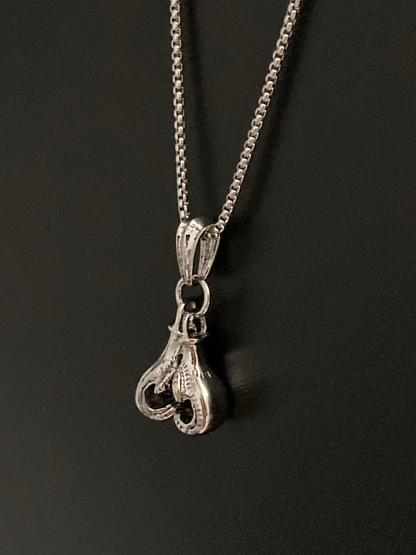 On a black background hangs a pair of boxing gloves stainless steel chain and pendant. Silver in colour this is a sturdy item that looks modern and is shiny in appearance. 