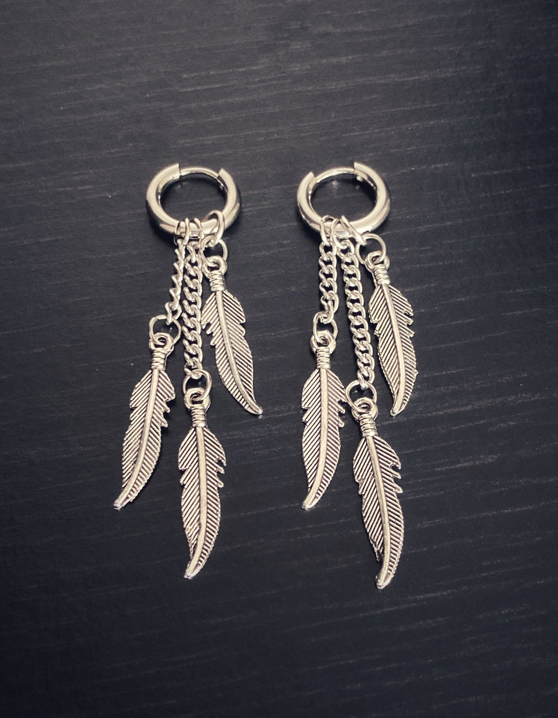 Lying next to each other are two small hoope earrings. Each with 3 feathers on chains hanging from the hoops themselves. All of the items are a shiny silver colour.