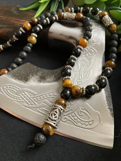 A stunning beaded necklace is draped over an axe. With a section that hangs down at the front and the rest of the item featuring metal cylindrical beads with patterning and also black shiny beads as well as tiger eye and black lava beads .