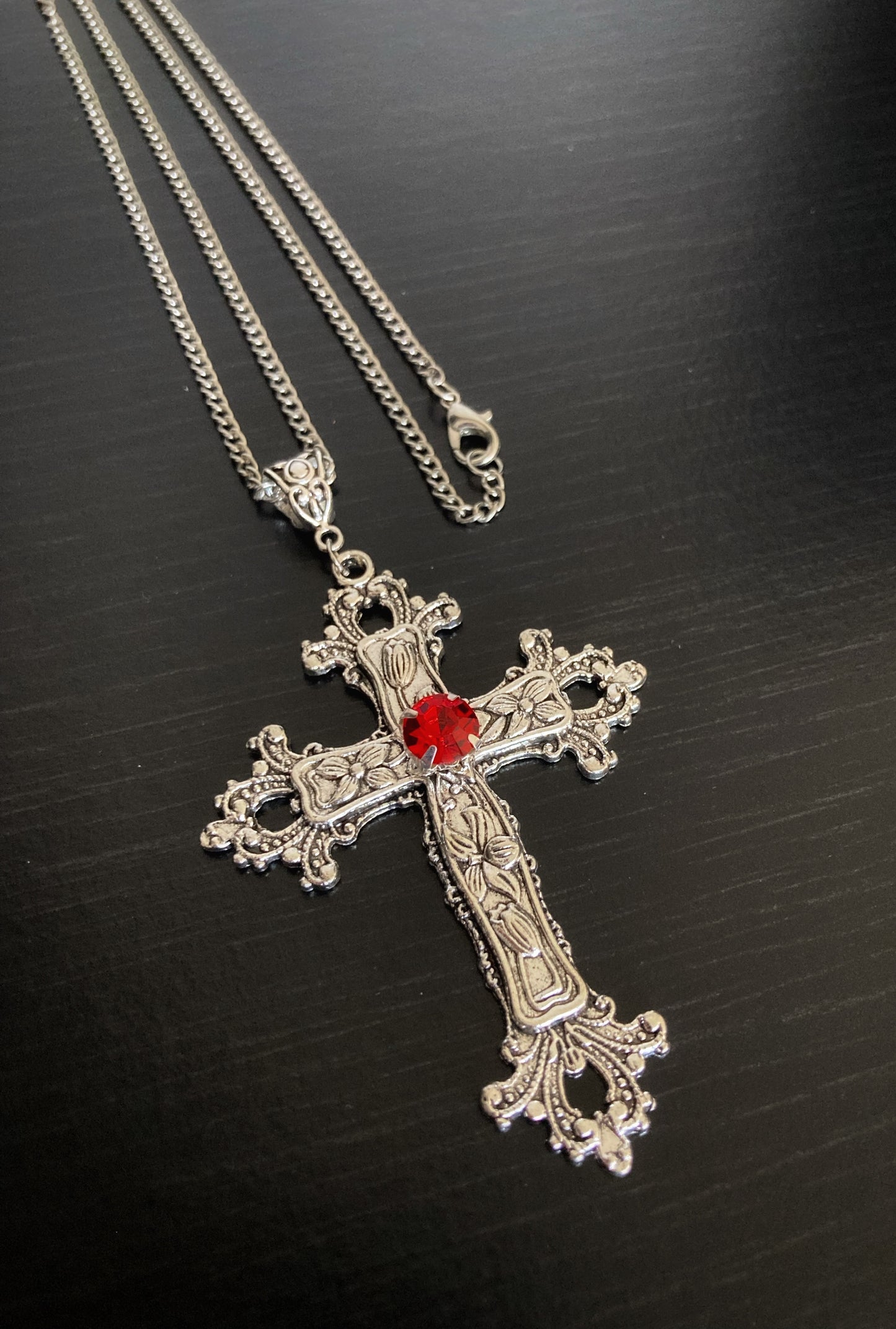 A full view of a silver coloured cross that looks gothic in style attached to a chain with a metal clasp. It is silver in colour apart from a red stone that is in the middle of the cross itself.