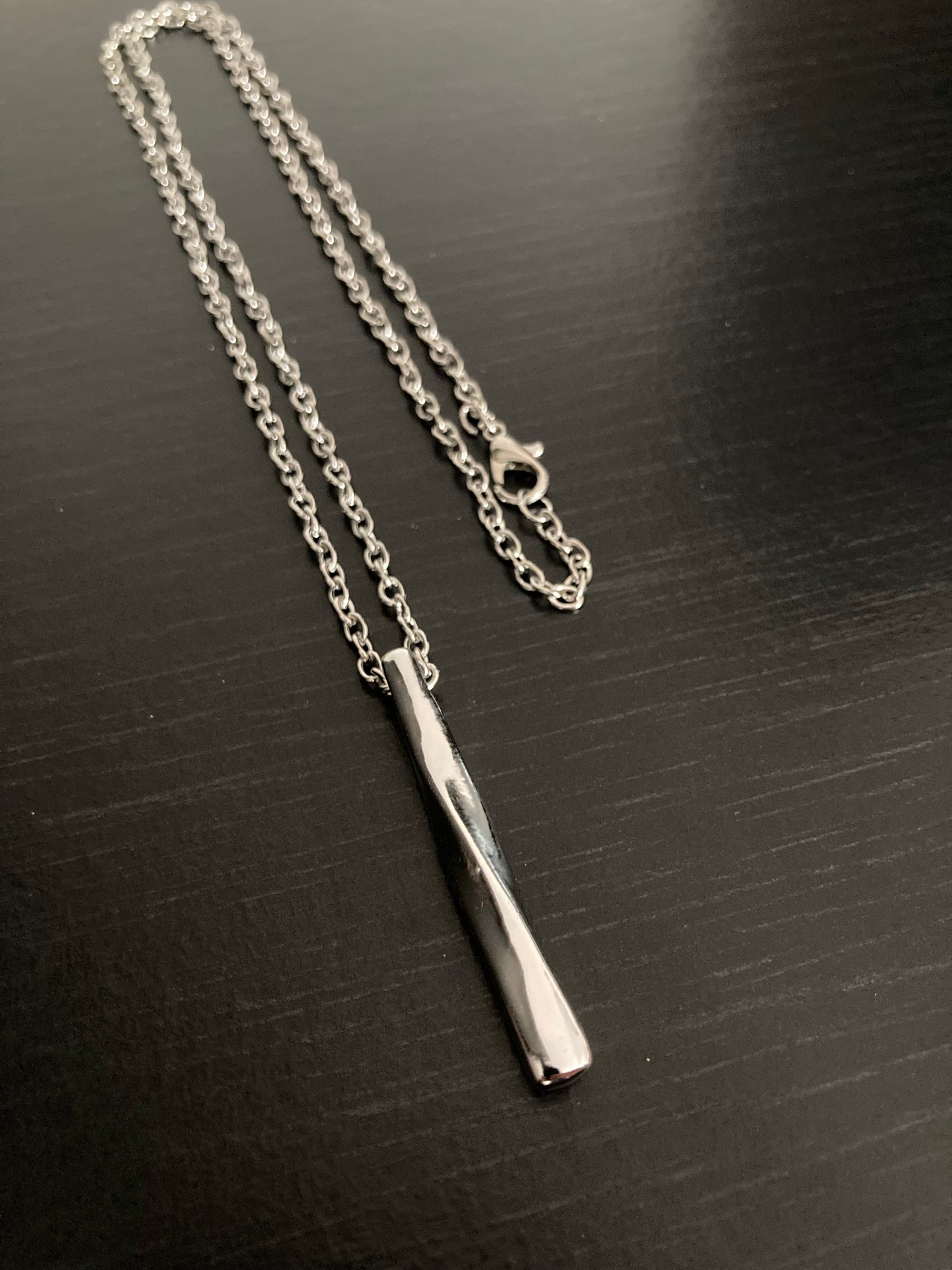 A silver coloured chain with a clasp for fastening lies on black surface. Attached to it is a pendant which is also silver and is a rectangular shape with a twisted design.