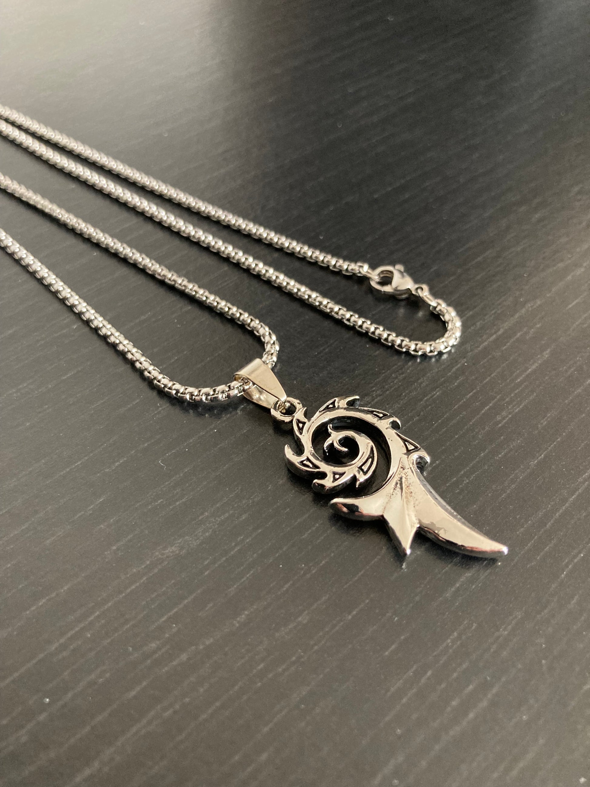 A black surface holds a silver coloured chain and pendant which is a spiral design. There is a metal clasp for fastening and the whole item is shiny. The spiral has a jagged look to it around the outer edges and it leads down to a spike at the end.