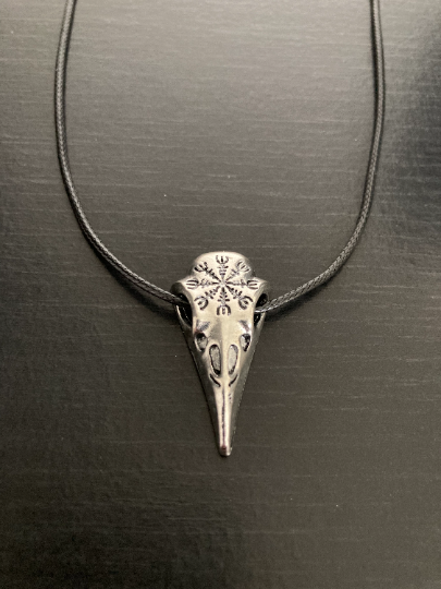 A front view of a raven skull pendant on a grainy black background featuring a black cord going through the eyes of the skull and a long beak with detailing on it. On the top of the skull itself is a engraving of the viking hekm of awe design.