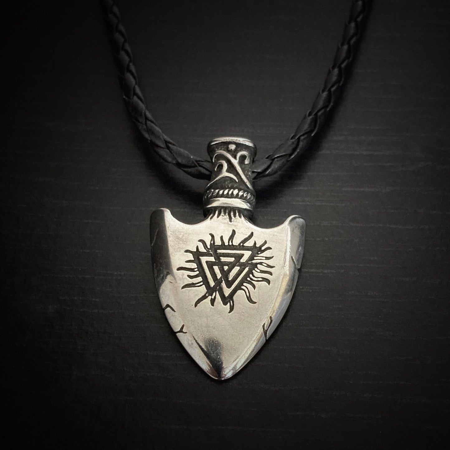 A silver coloured wide arrow head shaped pendant is hanging from a black cord. The design that can be seen is a valknut that hangs upside down. The cord goes through a patterned piece that has holes either side of it.