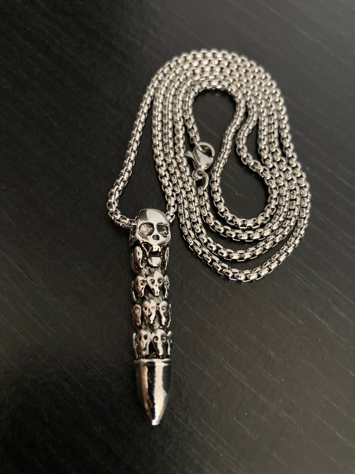 A silver colour chain is curled on a black surface with a three dimensional bullet shaped pendant attached to it. The quality metal clasp can be seen and the whole thing is made of a sturdy stainless steel. The bullet has skull heads carved onto it 