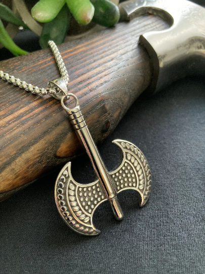 Propped against the handle of an axe is a silver coloured necklace with a two headed axe pendant. This is a detailed view and you are able to see the intricate detailing of the pattern on the item which you can also feel when you touch it.