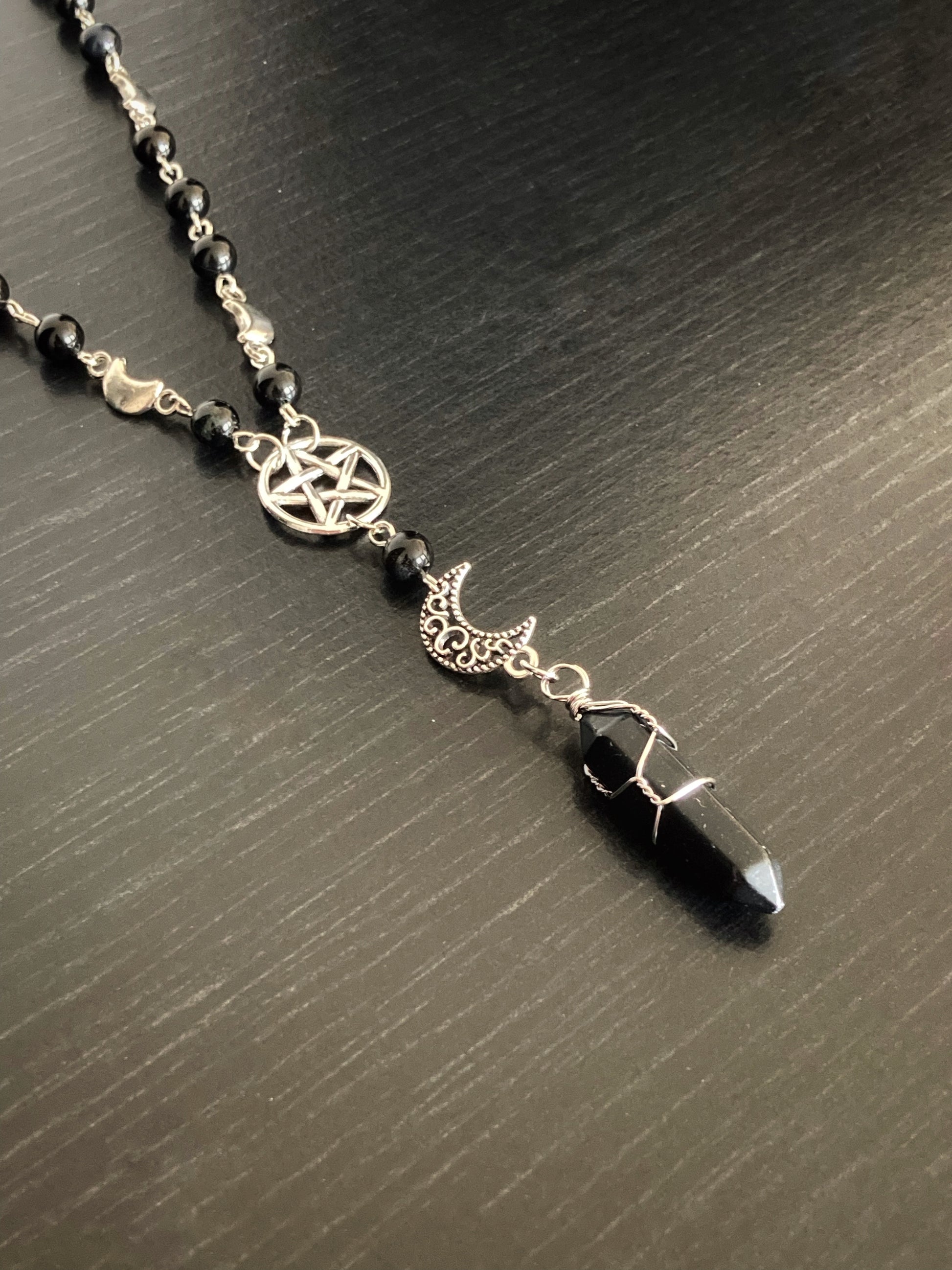 On a grey surface sits a section of a necklace. There is a pentagram in a circle with one black bead under it and then a patterned crescent moon. Below these is a onyx stone encased in a metal holder attaching it to the other items.