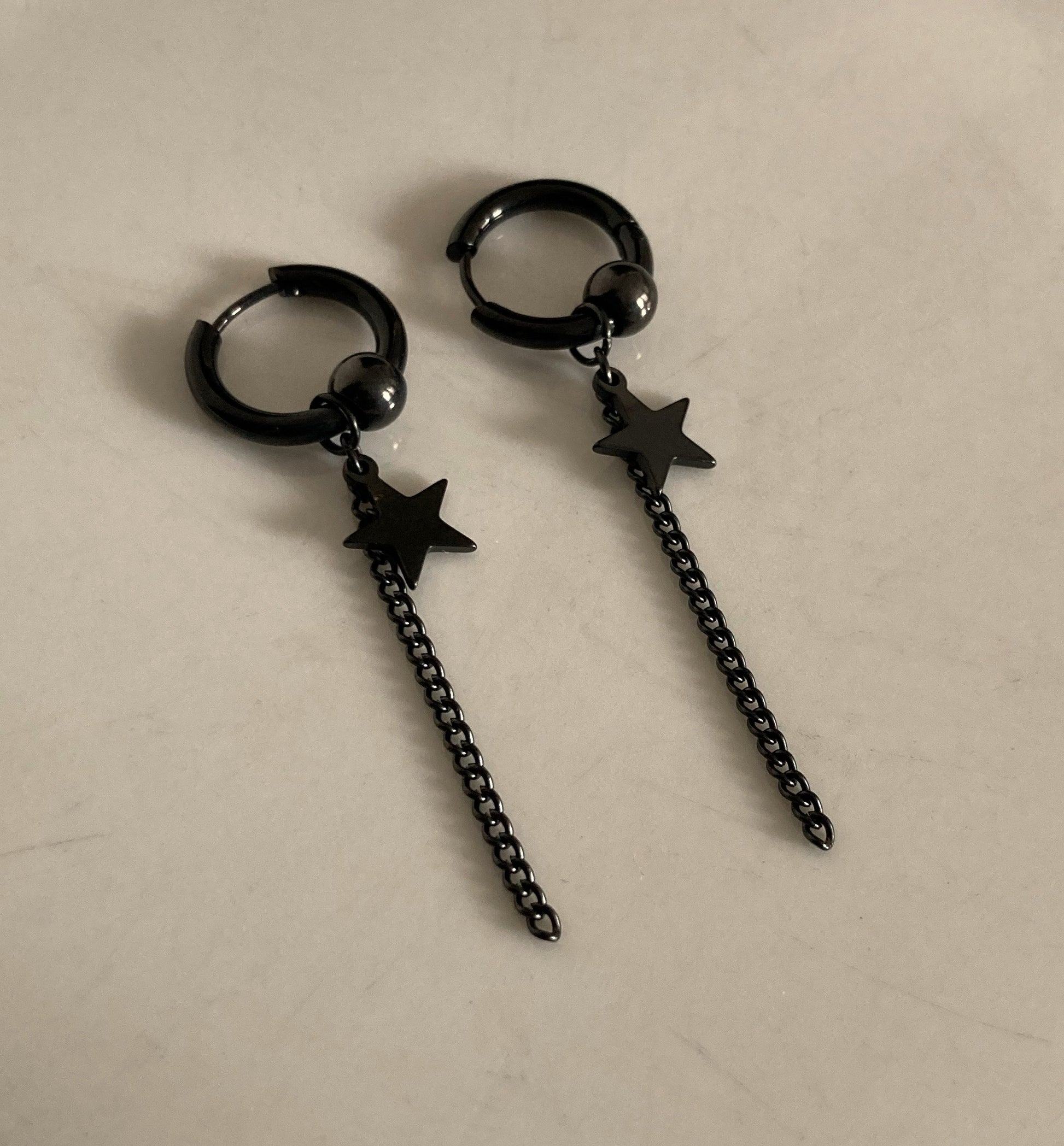 Two ring shaped earrings in black lie next to each other. A star hangs from each of them as does a chain. There is also a black solid round piece on them as well and a sleeper style bar that opens to put in the ear.